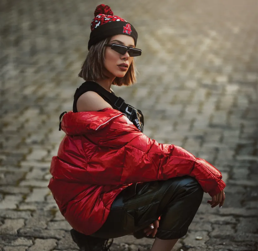 Stylish woman in red puffer jacket and beanie squatting on cobblestone.